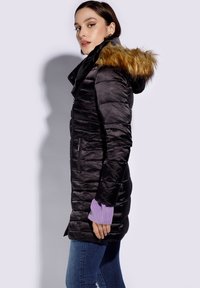 Abrigo acolchado negro con capucha de pelo sintético, bolsillos con cremallera y puños de canalé morados, que presenta un diseño ajustado y un texturizado acolchado.