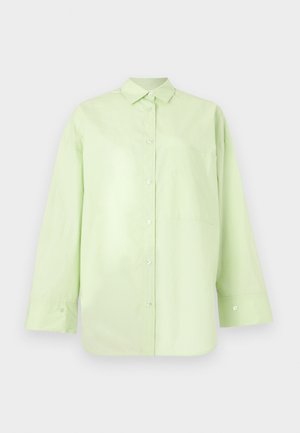 Femme aux longs cheveux noirs portant des lunettes transparentes, une chemise ample jaune clair boutonnée sur une robe marron foncé, et des chaussures plates foncées, debout à l'intérieur.