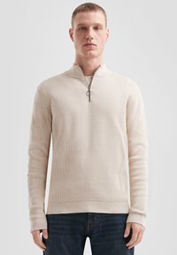 Beige zip-hals pullover trui met geribbelde textuur, lange mouwen en een zilveren rits. Draagt met donkere spijkerbroek tegen een neutrale achtergrond.