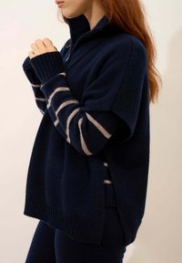 Granatowy sweter z suwakiem na kołnierzu, z beżowymi paskami na rękawach i ściągaczami. Miękka tekstura i luźny krój.