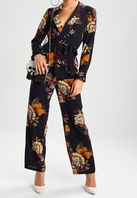 Completo a due pezzi blu navy con motivo floreale, vita incinturata, maniche lunghe e pantaloni a gamba larga, abbinato a una piccola borsa nera e tacchi.