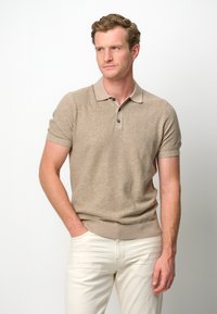 Beige short-sleeve polo shirt with a textured knit, two-button placket, and ribbed cuffs. Worn with light-colored slim-fit trousers.