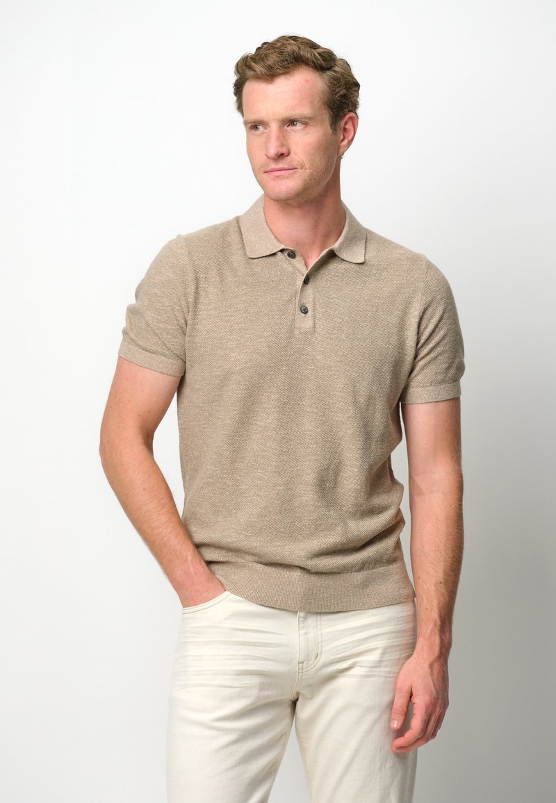 Beige short-sleeve polo shirt with a textured knit, two-button placket, and ribbed cuffs. Worn with light-colored slim-fit trousers.