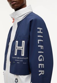 Μπλε ναυτικό και λευκό μπουφάν Tommy Hilfiger Marina Club με μεγάλο γράμμα «H» και κείμενο «Hilfiger» στο μανίκι που φοράει ένα άτομο.