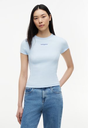 Kobieta ubrana w dopasowaną jasnoniebieską koszulkę z krótkim rękawem z napisem „tommy jeans” oraz wysokim stanem niebieskich dżinsów, stojąca na białym tle.