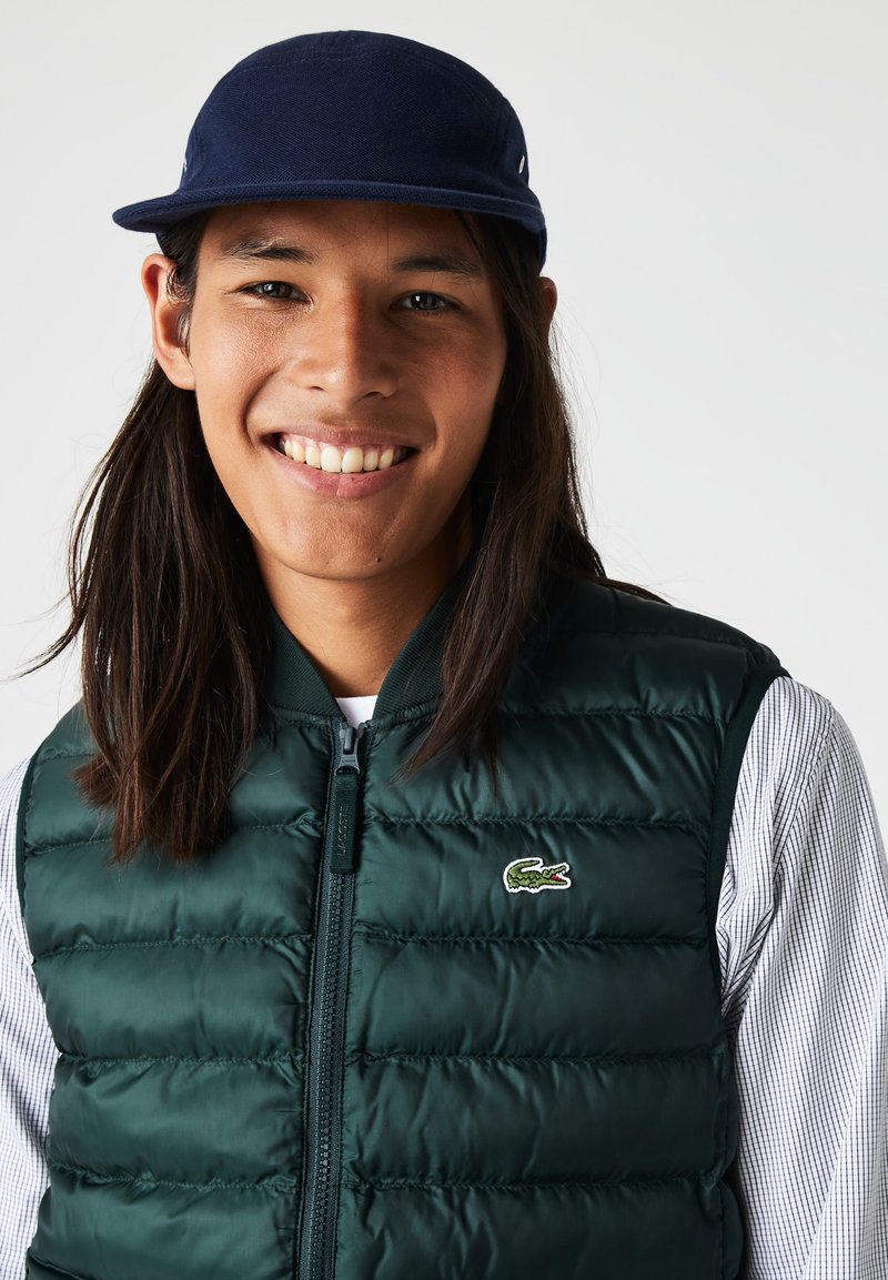 Persona joven sonriente con cabello largo y oscuro, usando una gorra azul marino, un chaleco acolchado verde con un logo de cocodrilo y una camisa de cuadros, sobre fondo blanco.