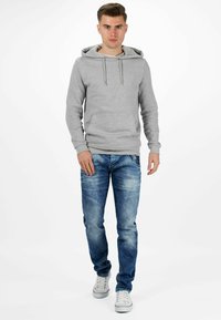 Haut à capuche gris avec cordon de serrage et poche, associé à un jean en denim bleu clair. Le modèle porte des baskets blanches, debout devant un fond blanc.