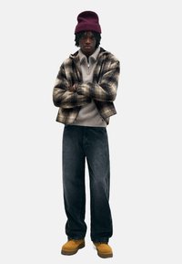 Rutigt jacka i bruna och krämiga nyanser, grå kordflorsbyxor och bruna stövlar. Bärs med en vinröd stickad mössa och en krämfärgad ziptröja.