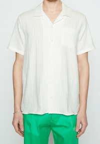 Camicia bianca a maniche corte con tessuto testurizzato. Caratterizzata da una tasca sul lato sinistro del petto e un colletto classico. Abbinata a pantaloni verdi.