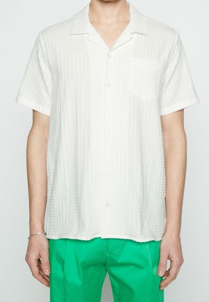 Chemise blanche à manches courtes, boutonnée, avec un tissu texturé. Comprend une poche sur la poitrine gauche et un col classique. Associée à un pantalon vert.