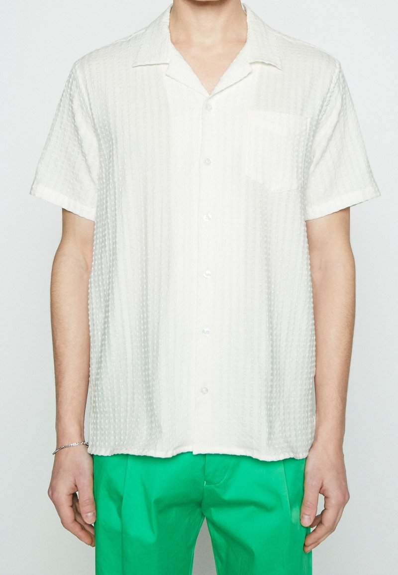 Camicia bianca a maniche corte con tessuto testurizzato. Caratterizzata da una tasca sul lato sinistro del petto e un colletto classico. Abbinata a pantaloni verdi.