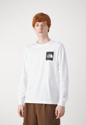 Camiseta blanca de manga larga hecha de algodón, que cuenta con un logo cuadrado negro en el pecho izquierdo. El modelo lleva pantalones cortos cargo marrones.