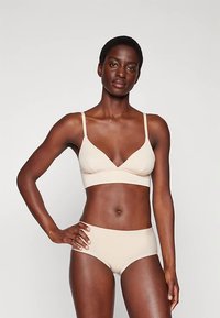 Žena s krátkými vlasy, oblečená v béžovém prádle a odpovídajících vysokých kalhotkách, stojící s jednou rukou na boku na neutrálním pozadí.