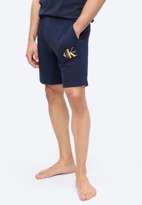 Mann iført marineblå Calvin Klein-shorts med lommer, stående barbeint mot en hvit bakgrunn.