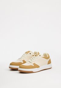 Zapatillas de deporte de piel sintética en beige y marrón claro, con una suela de goma blanca, diseño de cordones y detalles texturizados en los laterales.