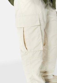 Beige cargo pants with multiple side pockets, made of lightweight, textured fabric. Features a loose fit and subtle stitching details.