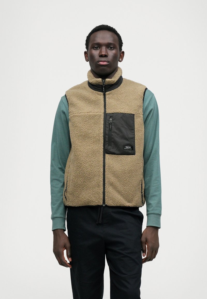 Καφέ γιλέκο από φλις με ψηλό γιακά, μαύρη τσέπη με φερμουάρ στο στήθος, αντίθεση πράσινης μπλούζας με μακριά μανίκια από κάτω, φορεμένο με μαύρο παντελόνι.