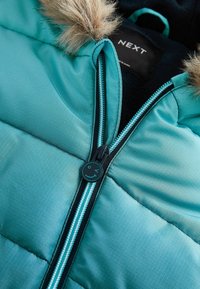 Chaqueta acolchada de color azul verdoso con un ribete de piel en la capucha, con el zipper parcialmente desabrochado que presenta un tirador con una carita sonriente.