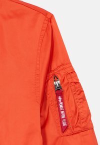 Rękaw pomarańczowej kurtki z zasuwaną kieszenią z tagiem "USUŃ PRZED LOTEM", wykonany z gładkiego materiału z wzmocnionymi szwami.