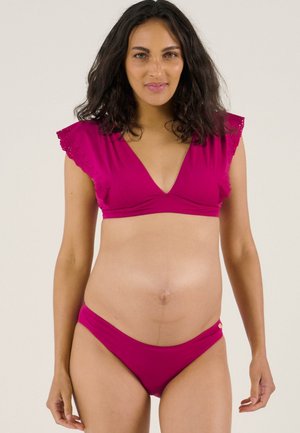 Schwangere Frau mit schulterlangem dunklem Haar, die einen magentafarbenen Bikini mit gerüschten Cap-Ärmeln trägt und vor einem einfarbigen Hintergrund steht.