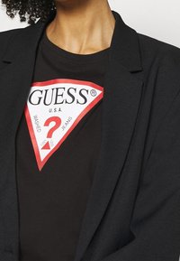 Camiseta de algodón negra con un logo triangular en rojo y blanco que lleva el texto "GUESS U.S.A." Una chaqueta de un tejido negro estructurado cubre los hombros.