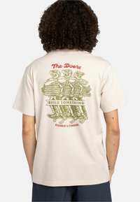 Krémovobiele tričko s krátkym rukávom s farebnou grafikou troch kostier na chrbte, ktoré držia polená, a výrazným textom nad nimi.