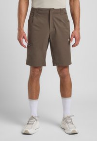 Pantaloni corti marroni con una texture liscia, lunghezza al ginocchio, dotati di tasche laterali e dettagli con zip. Indossati con calze bianche e sneakers di colore chiaro.