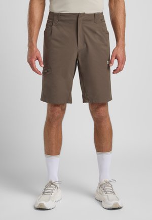 Shorts marron avec une texture lisse, arrivant au genou, avec des poches latérales et un détail de fermeture éclair. Portés avec des chaussettes blanches et des baskets claires.