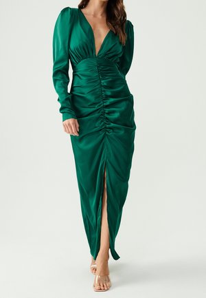 Abito in raso verde con scollatura a V profonda, maniche lunghe, parte anteriore arricciata e spacco alto fino alla coscia. Tessuto liscio e silhouette aderente. Tacchi trasparenti.