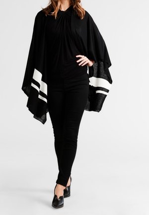 Frau trägt schwarze Hose und Oberteil mit einem schwarz-weiß gestreiften Schal, steht mit einer Hand in der Hüfte und überschlagenen Beinen.