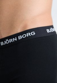 Taille und Hüfte einer Person, die schwarze Björn Borg Unterwäsche mit einem sichtbaren, markierten elastischen Bund trägt.