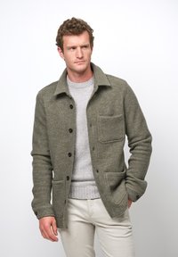 Olivgrön ullblandad jacka med krage, framknäppning, bröstficka och dubbla nedre fickor, som bärs över en ljusgrå tröja.