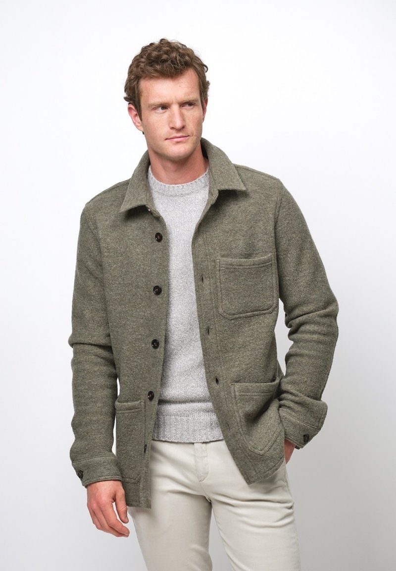Olivgrön ullblandad jacka med krage, framknäppning, bröstficka och dubbla nedre fickor, som bärs över en ljusgrå tröja.