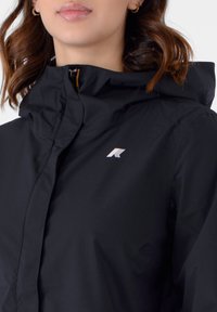 Giacca nera impermeabile con una leggera lucentezza, dotata di collo con zip, cappuccio e un piccolo logo bianco sul lato sinistro del petto.