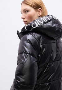 Μαύρο μπουφάν τύπου puffer με γυαλιστερό φινίρισμα, oversized κουκούλα και λευκό λογότυπο "Calvin Klein" εμφανώς τοποθετημένο στον γιακά.