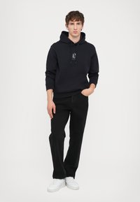 Marineblaue Kapuzenjacke mit Fronttasche und Logo. Trägt schwarze Jeans und weiße Sneakers, steht vor einem schlichten Hintergrund.