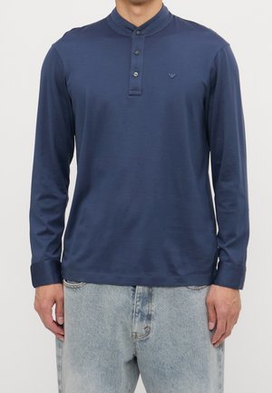 Man draagt een marineblauw henley-shirt met lange mouwen en drie knopen en lichtblauwe verweerde jeans, staand tegen een effen achtergrond.