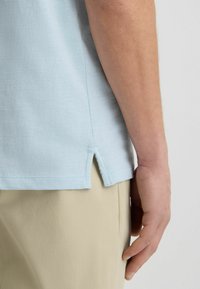 T-shirt azzurro chiaro con scollatura rotonda, maniche corte e spacchetti laterali. Tessuto morbido e dettaglio di orlo cucito. Indossato con pantaloni beige.