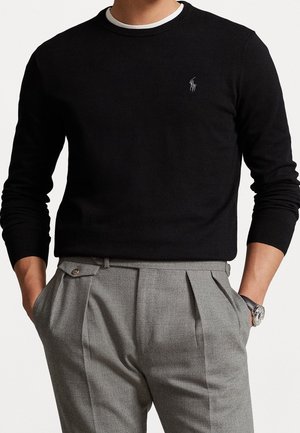 Mężczyzna ubrany w czarny sweter z długim rękawem z białym obszyciem oraz szare plisowane spodnie, ręce w kieszeniach, zegarek na lewej ręce.