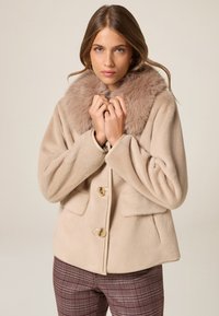 Abrigo de piel sintética beige con un amplio cuello, cierre de botones y dos bolsillos frontales. Textura suave y diseño amplio; llevado sobre pantalones de cuadros.