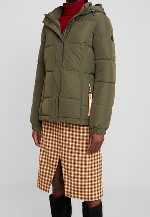 Osoba nosící olivově zelenou péřovou bundu s kapucí, červený rolák, hnědou sukni s psím zubem a předním rozparkem a černé kozačky.