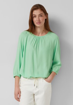 Jeune femme aux longs cheveux bruns portant un chemisier ample vert menthe et un pantalon blanc, se tenant devant un fond gris uni.