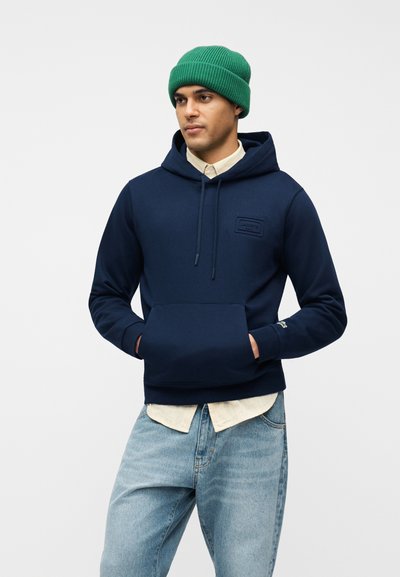 Homme portant un bonnet vert, un sweat à capuche bleu marine avec poche avant, une chemise beige boutonnée et un jean bleu clair, debout devant un fond blanc.