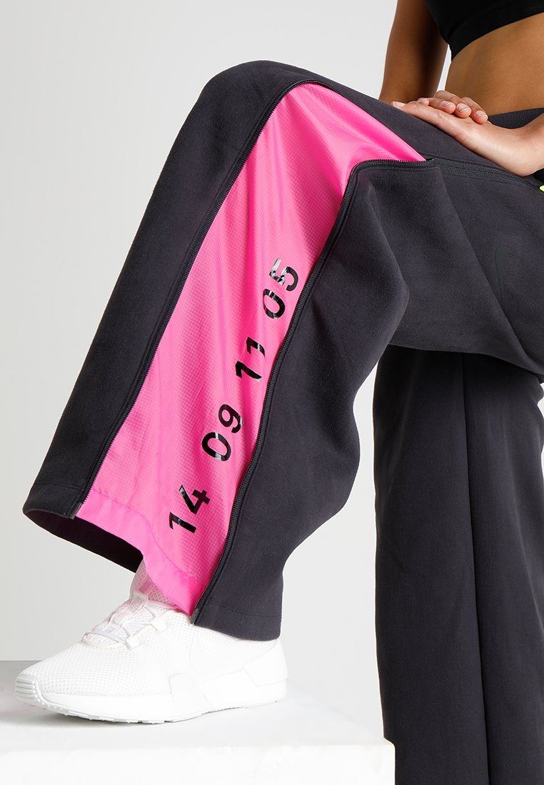 Pantalon large noir avec un panneau en maille rose, affichant les numéros imprimés "14 09 11 05." Le matériau semble doux ; accompagné de chaussures de sport blanches.