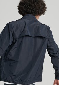 Chaqueta ligera de color azul marino con una textura suave, dobladillo elástico y un detalle de solapa en la parte trasera. Presenta un cuello clásico y mangas largas.