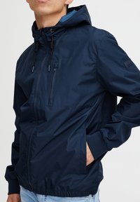 Tummansininen huppari, joka on valmistettu sileästä, kevyestä kankaasta, jossa on joustavat hihansuut, vetoketjullinen rintatasku ja säädettävät nyörikiinnitykset.