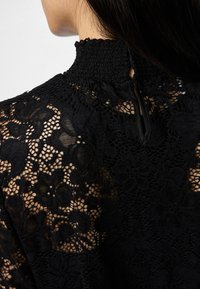 Blusa de renda preta com gola alta pregueada, padrões florais e um design ornamentado com textura transparente. Fecho em forma de chave nas costas.
