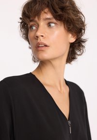 Czarny top zapinany na zamek z dekoltem w kształcie litery V, wykonany z gładkiego materiału. Charakteryzuje się dopasowanym krojem i minimalistycznym stylem, podkreślającym czyste linie.