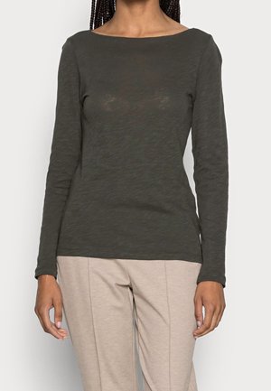 Kvinde iført en mørkegrøn, tætsiddende, langærmet top og lysebeige bukser, stående med armene afslappet ned langs siderne mod en ensfarvet baggrund.