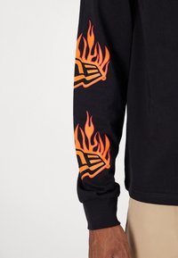 Langarmshirt in Schwarz mit orangenen Flammendrucken auf beiden Ärmeln und einem Logodesign am Oberarm. Weicher Baumwollstoff.